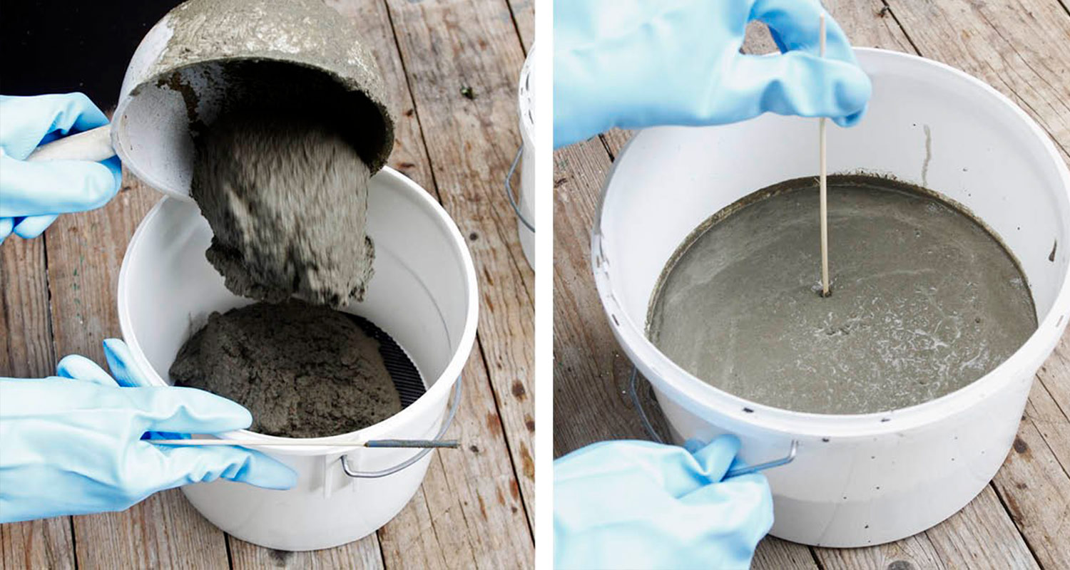 Siniset hanskat kaatavat ja sekoittavat harmaata betonimassaa valkoisessa ämpärissä puupinnalla
