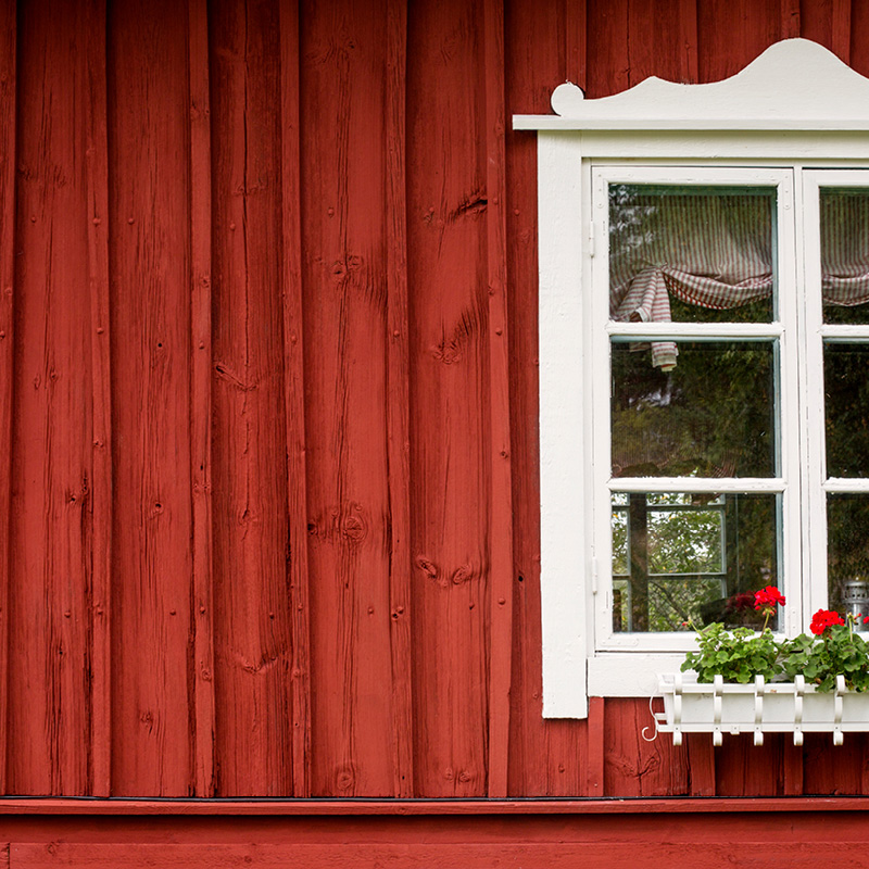 Punaisen puuverhouksen keskellä valkoinen ikkuna, pelargonit ikkunalaudalla