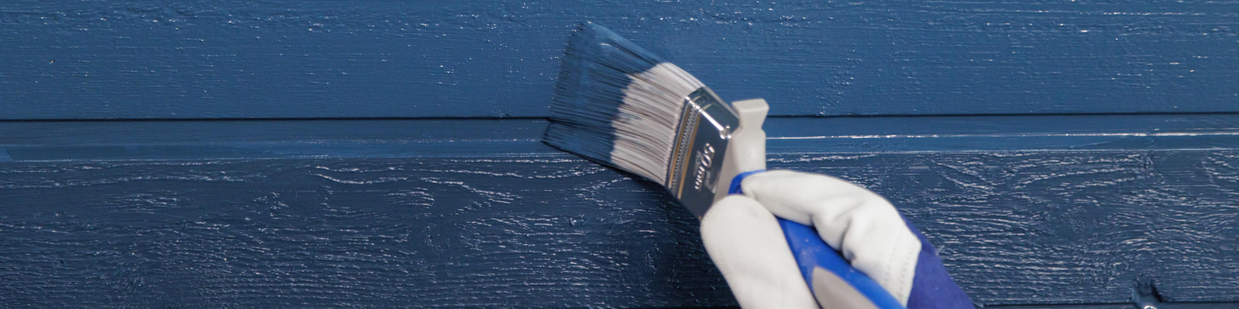 Käsineellinen käsi siveltimellä maalaa puupaneelin ulkoseinää tummansiniseksi.