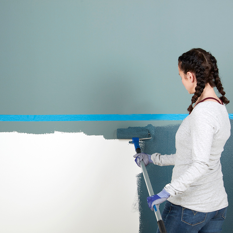 Nainen maalaa rullalla sisäseinän sinertävällä maalilla valkoisen pohjan päälle.