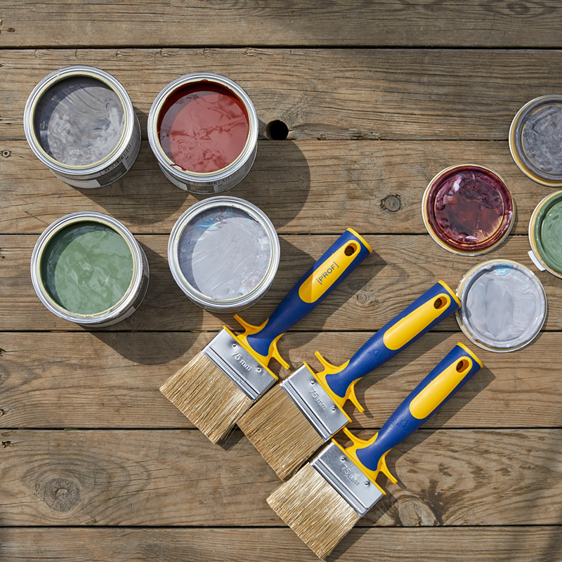 Avoimet maalipurkit eri väreissä ja kolme sinikeltakahvaista sivellintä puupöydällä