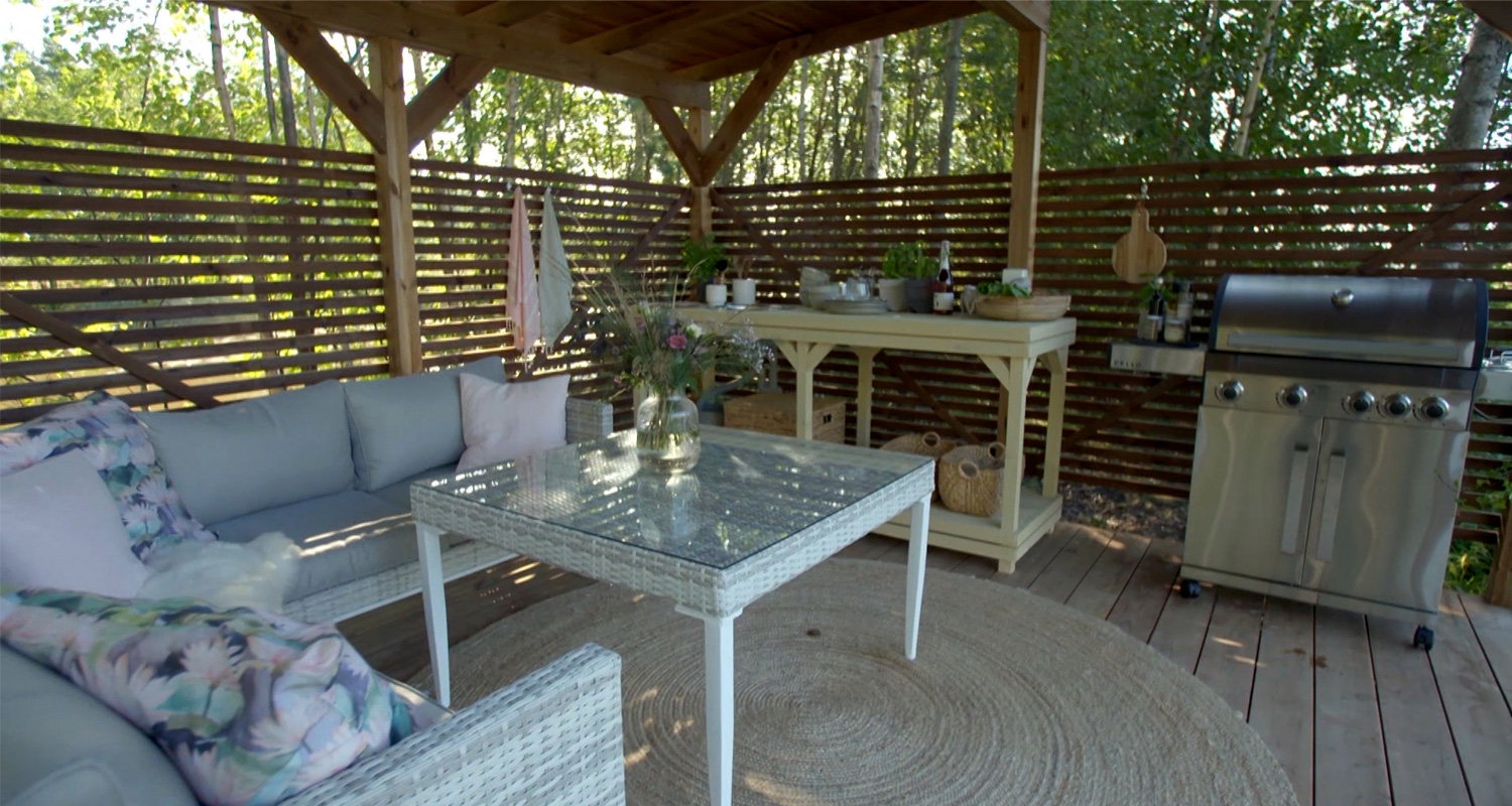 Katettu terassi metsässä, vaalea punossohva pastellityynyin, lasipöytä juuttimatolla ja grilli