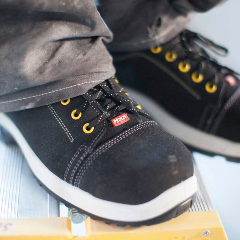 Musta suojakenkä keltaisilla nauhanreijillä ja valkoisella pohjalla metallitikasaskelmalla.