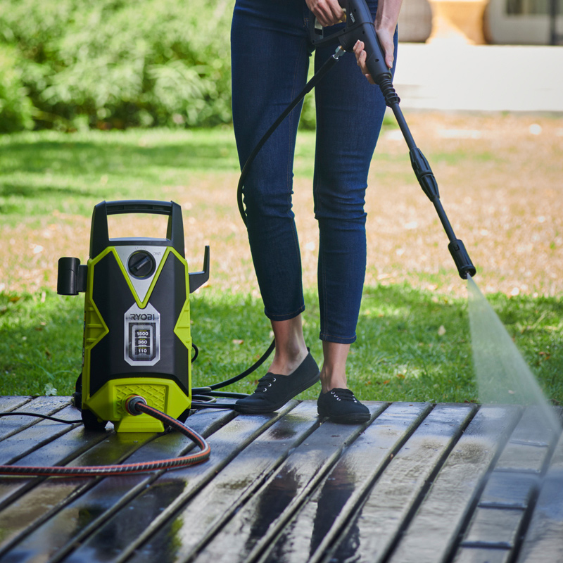Farkkuhousuinen henkilö pesee vesisuihkulla vihreä-mustalla painepesurilla puuterassia