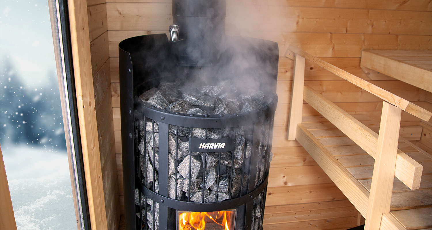 Mustan Harvia-kiukaan höyryävät kivet puusaunassa, lauteet oikealla ja luminen metsä ovesta.