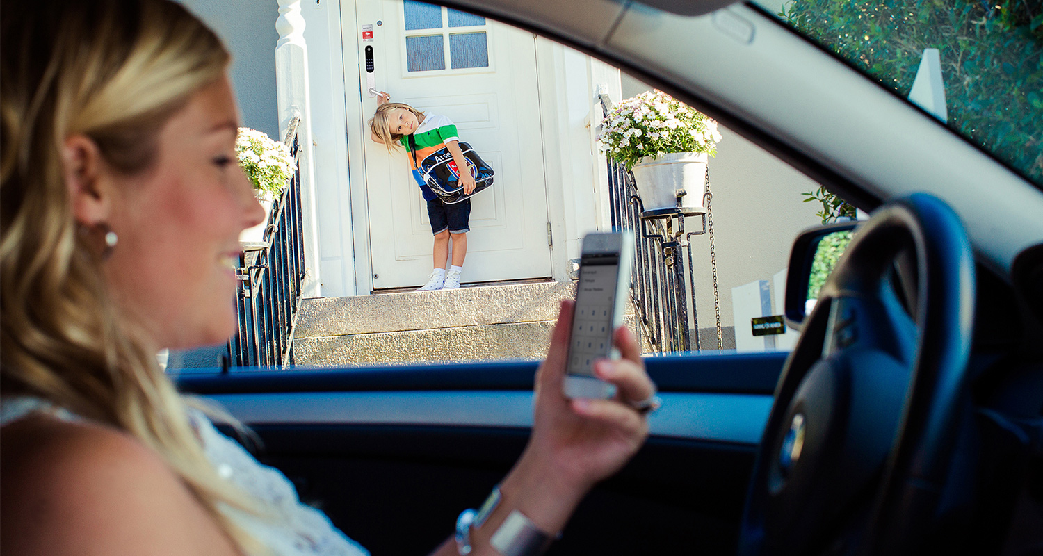 Nainen autossa kännykkä kädessä, portailla lapsi repun kanssa valkoisen talon edessä