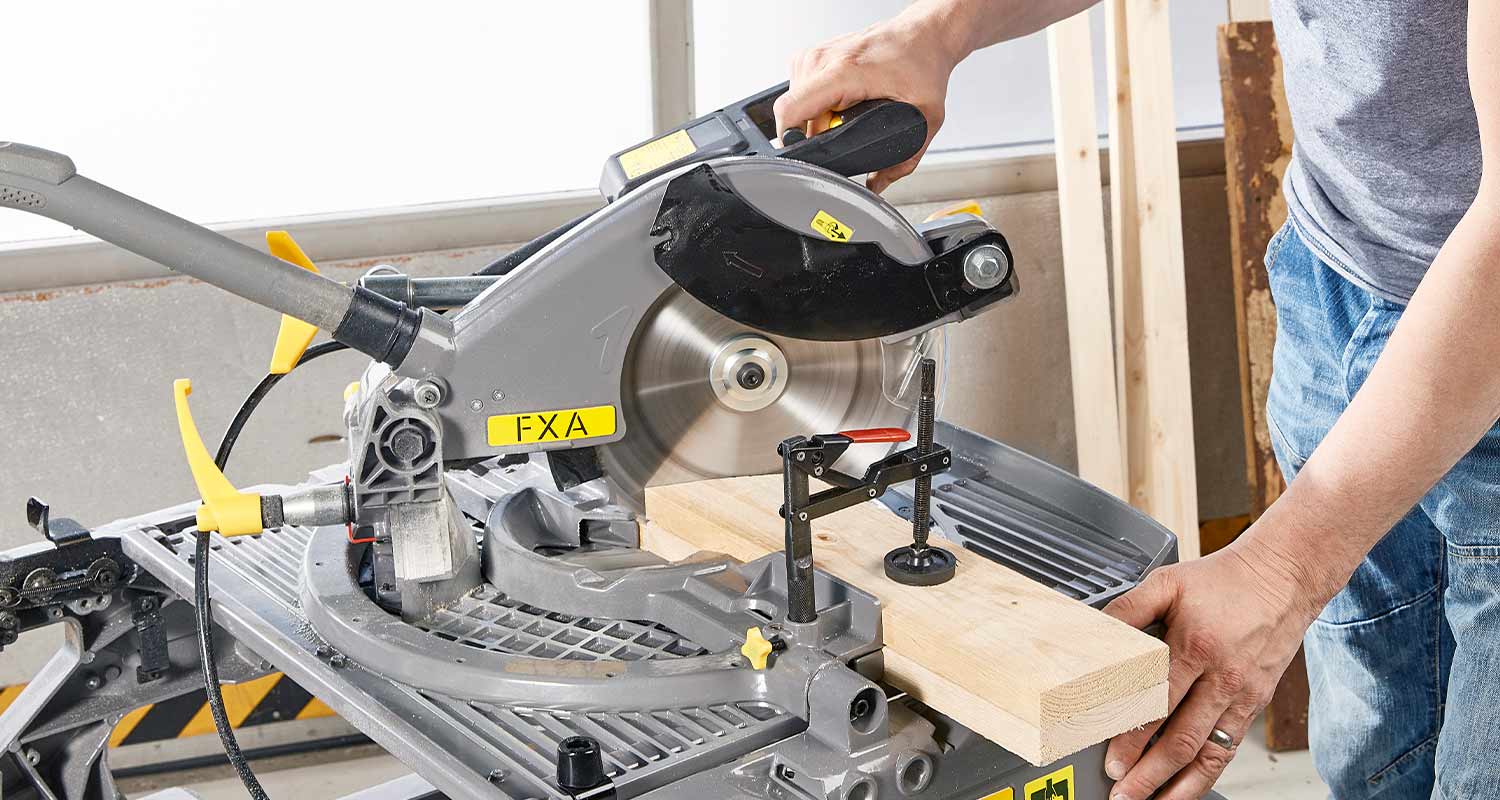 Harmaa-keltainen jiirisaha leikkaa puupalkkia verstaassa, kädet ohjaavat sahausta.