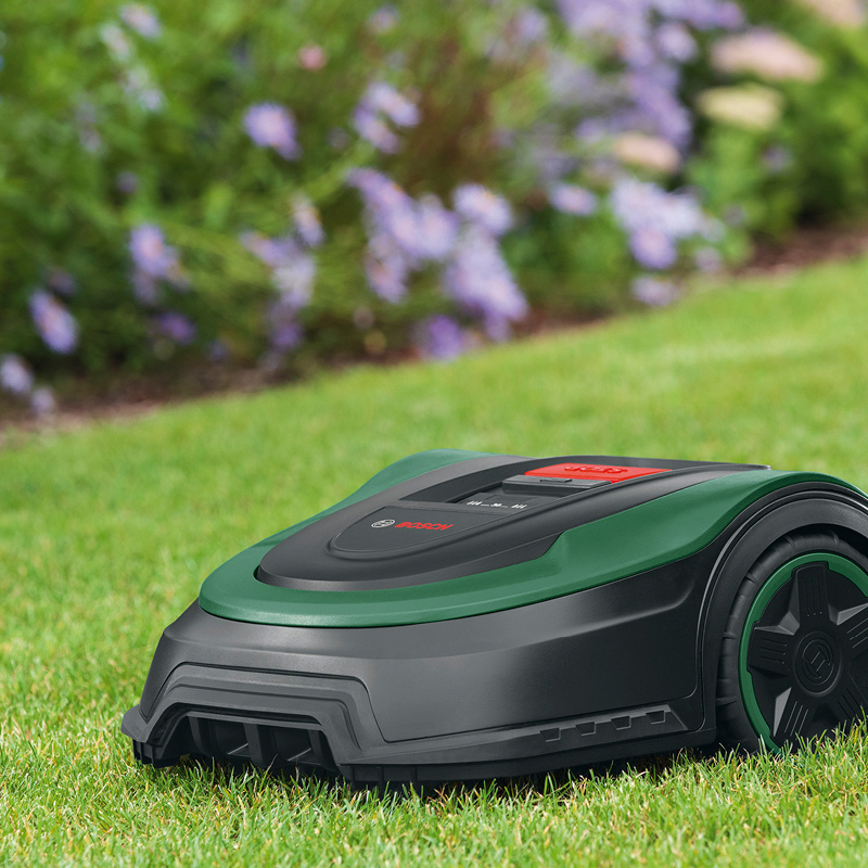 Vihreä-musta Bosch-robottiruohonleikkuri nurmikolla, lilakukkapensaat taustalla.