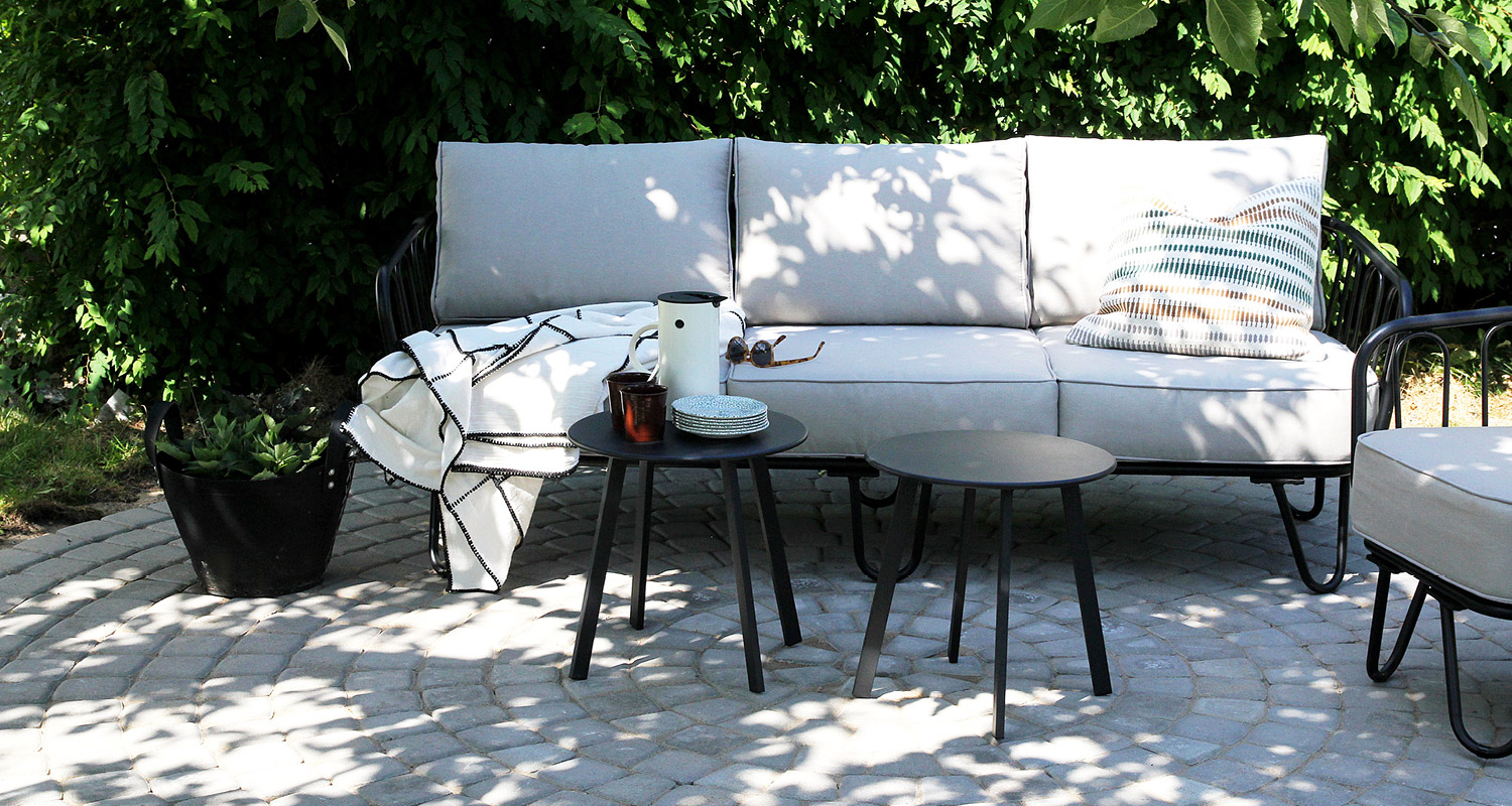 Harmaa ulkosohva mustilla jaloilla, kaksi mustaa pyöröpöytää astioin, vihreä aita taustalla.