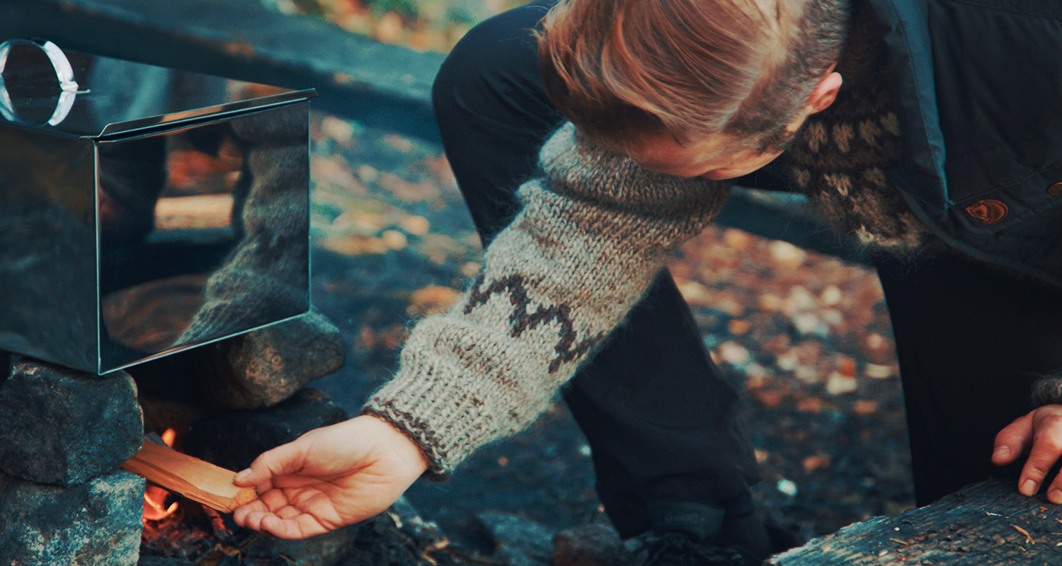 Harmaavillapuseroinen henkilö lisää puuklapia hopeiselle retkikeittimelle syksyisessä metsässä
