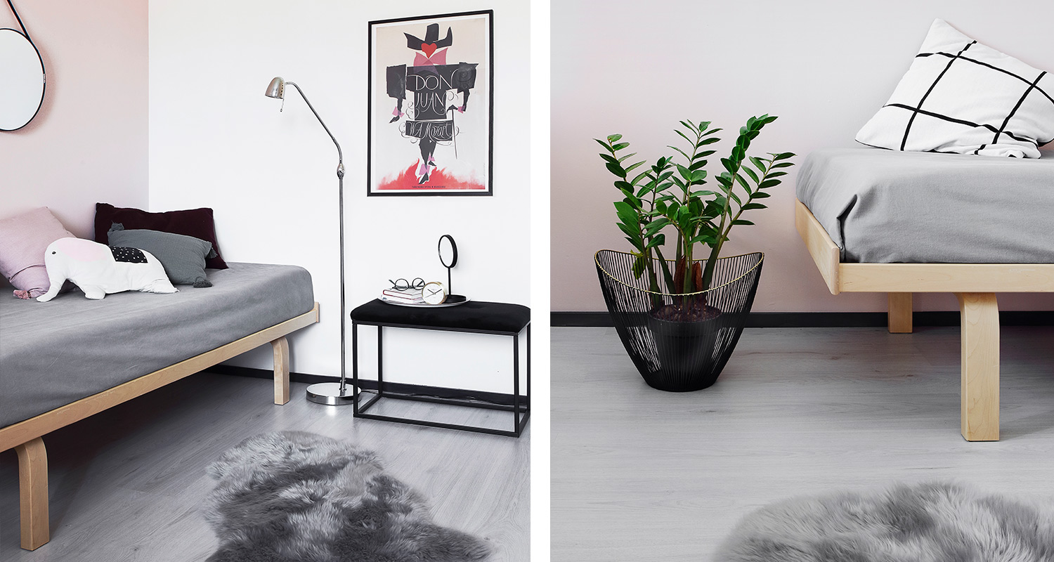 Vaaleanpunainen huone: puiset sängyt harmailla peitteillä, musta penkki, viherkasvi ja juliste