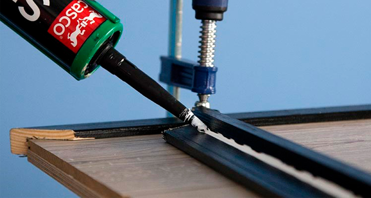 Vihreä-musta tiivistysmassaputki puristaa valkoista massaa mustaan kehykseen puisella alustalla.