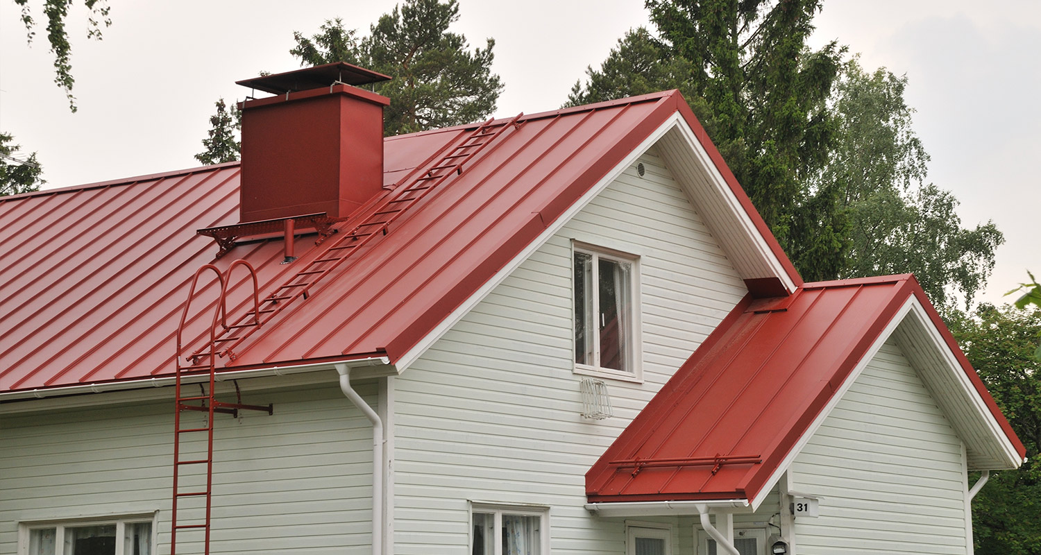 Valkoinen puutalo punaisella peltikatolla, tikkaat ja piippu, taustalla havumetsää