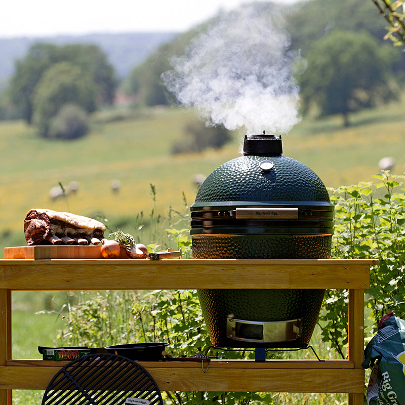 Vihreä savustinuuni savuaa puupöydällä, vieressä paistiliha ja sipulit, niitty taustalla.