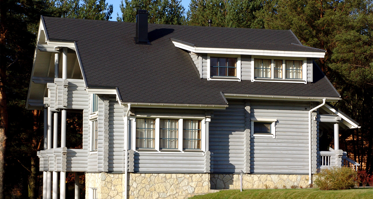 Vaaleanharmaa hirsitalo mustalla katolla ja kivijalalla mäntymetsän edessä