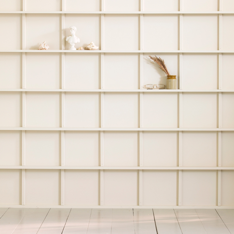 Valkoinen ruutuhyllyseinä, jossa pieni patsas, simpukoita, purkki kuivia oksia ja silmälasit.