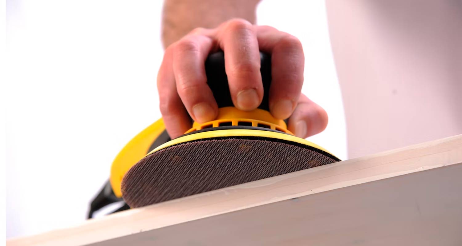 Käsi pitelee kelta-mustaa pyöröhiomakonetta hioen vaaleaa puupintaa.