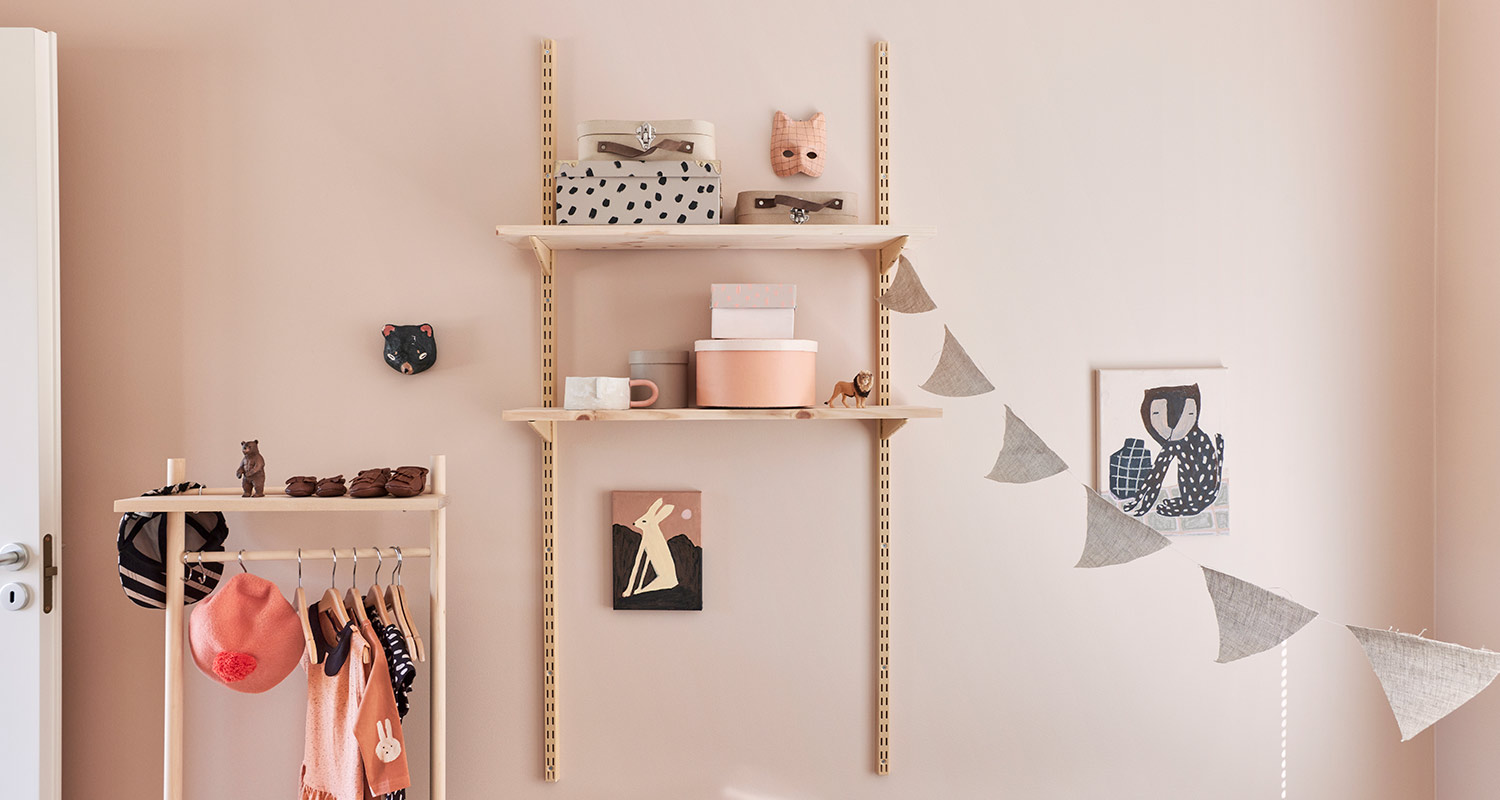 Lastenhuoneen vaaleanpunainen seinä, puuhyllyt, vaateripustin ja viirikoriste.