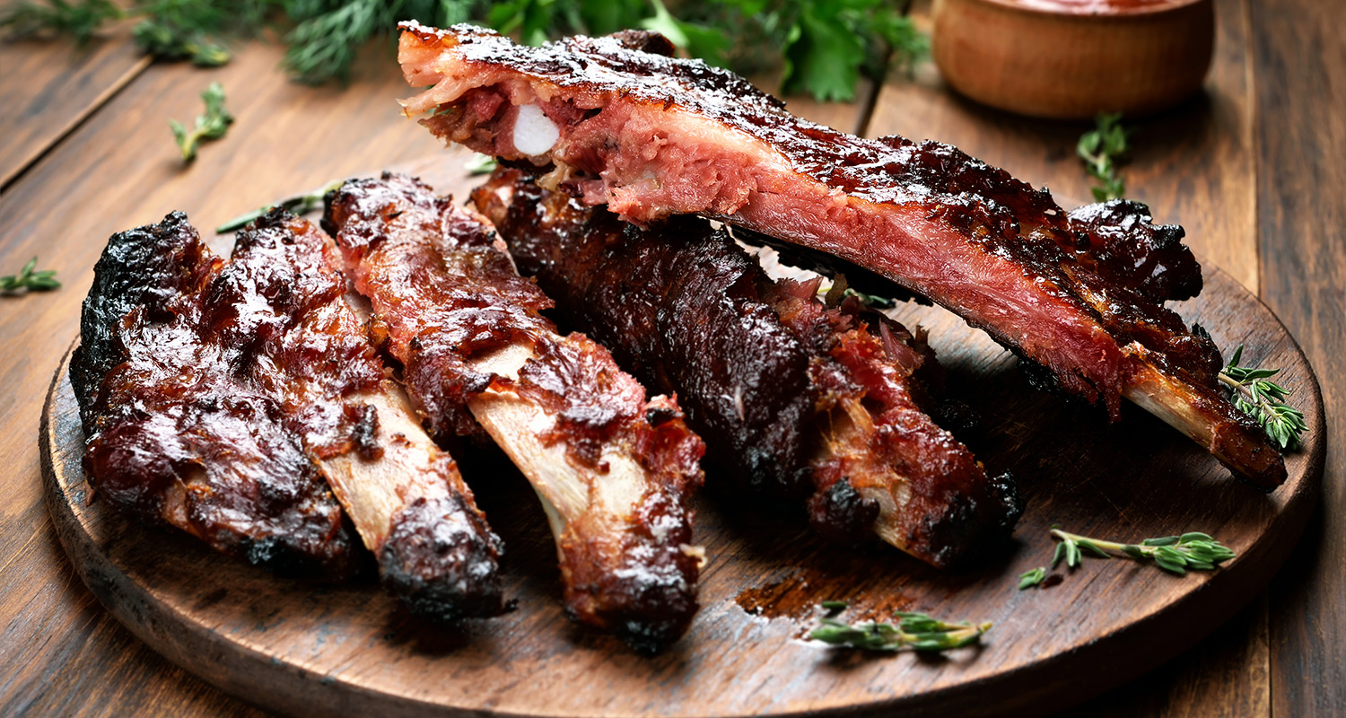 Savustettuja BBQ-kylkipaloja tumman laudan päällä rosmariinin oksien kera