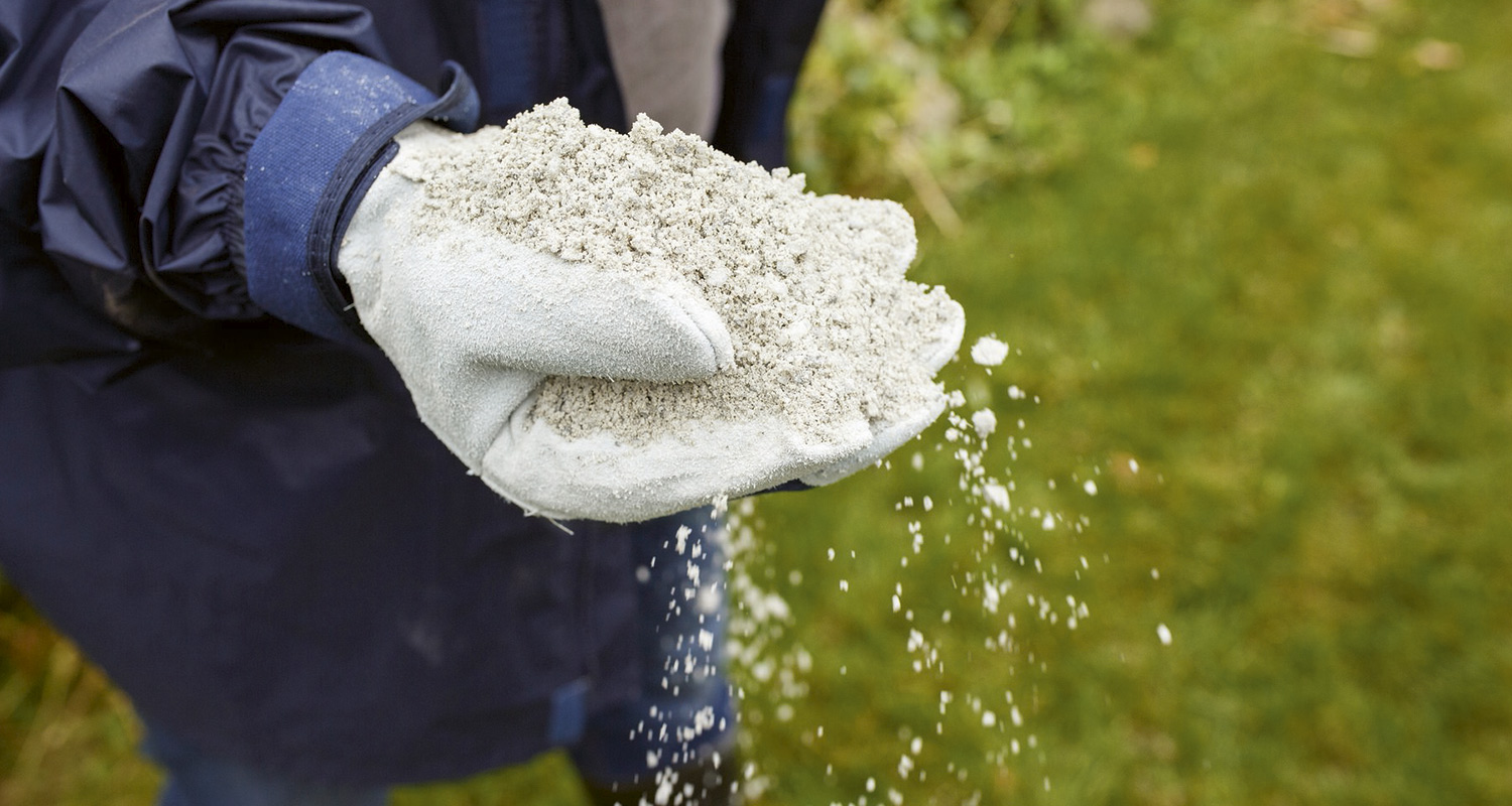 Työkäsineiset kädet pitelevät harmaita rakeita, jotka valuvat vihreälle nurmelle.