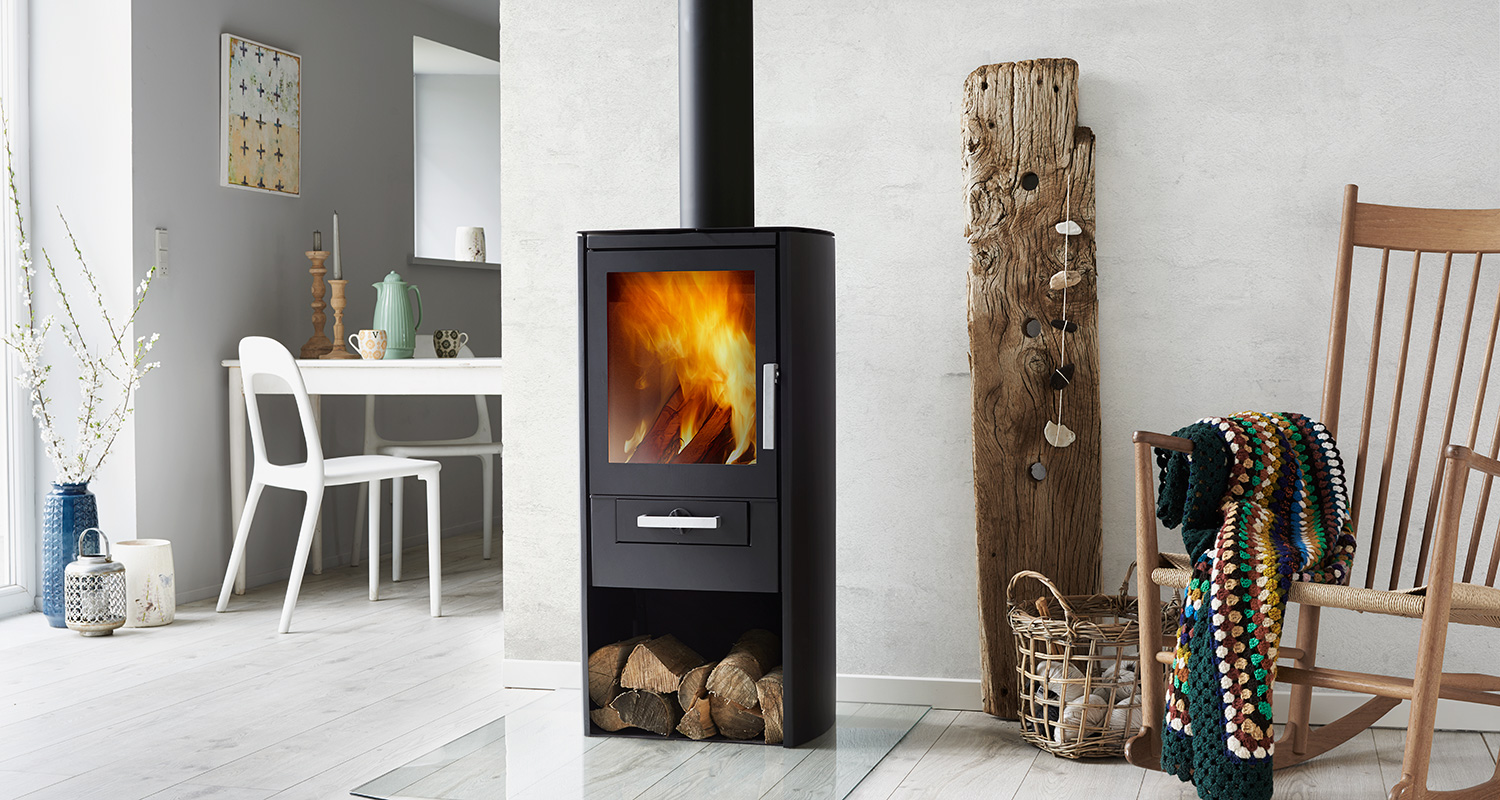 Musta takka tulenloimussa, polttopuita, värikäs virkattu viltti keinutuolissa ja vaalea sisustus