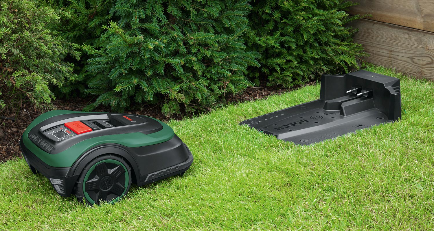 Vihreä-musta robottiruohonleikkuri ja sen musta latausasema nurmella kuusiaidan edessä