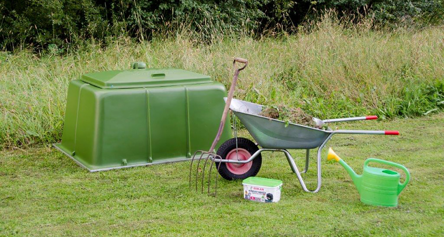 Vihreä kompostisäiliö pihanurmella, edessä kottikärryt ruohosilpulla, harava ja kastelukannu.