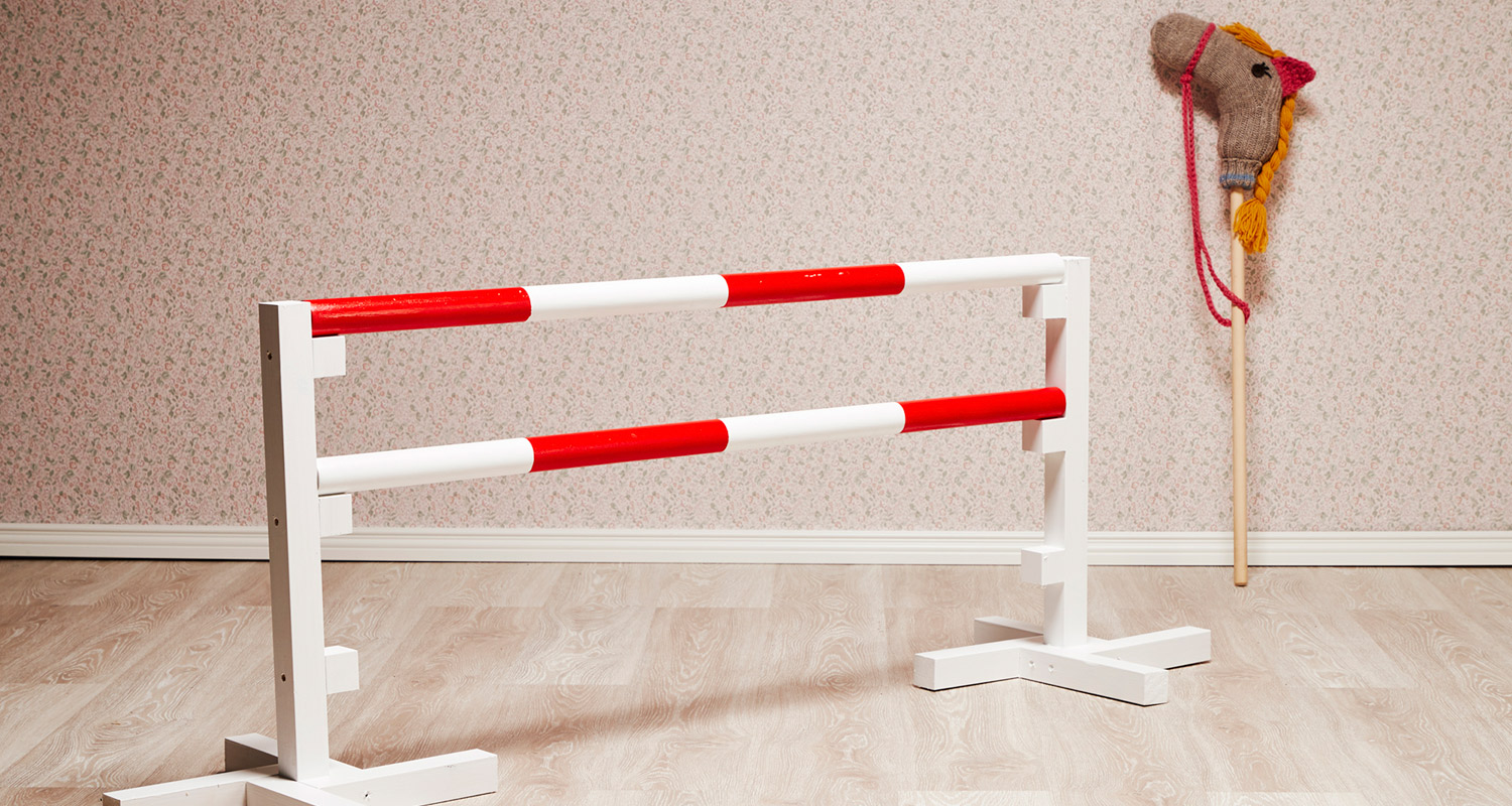 Valko-punainen ratsastuseste puulattialla kukkaseinää vasten, seinässä virkattu keppihevonen