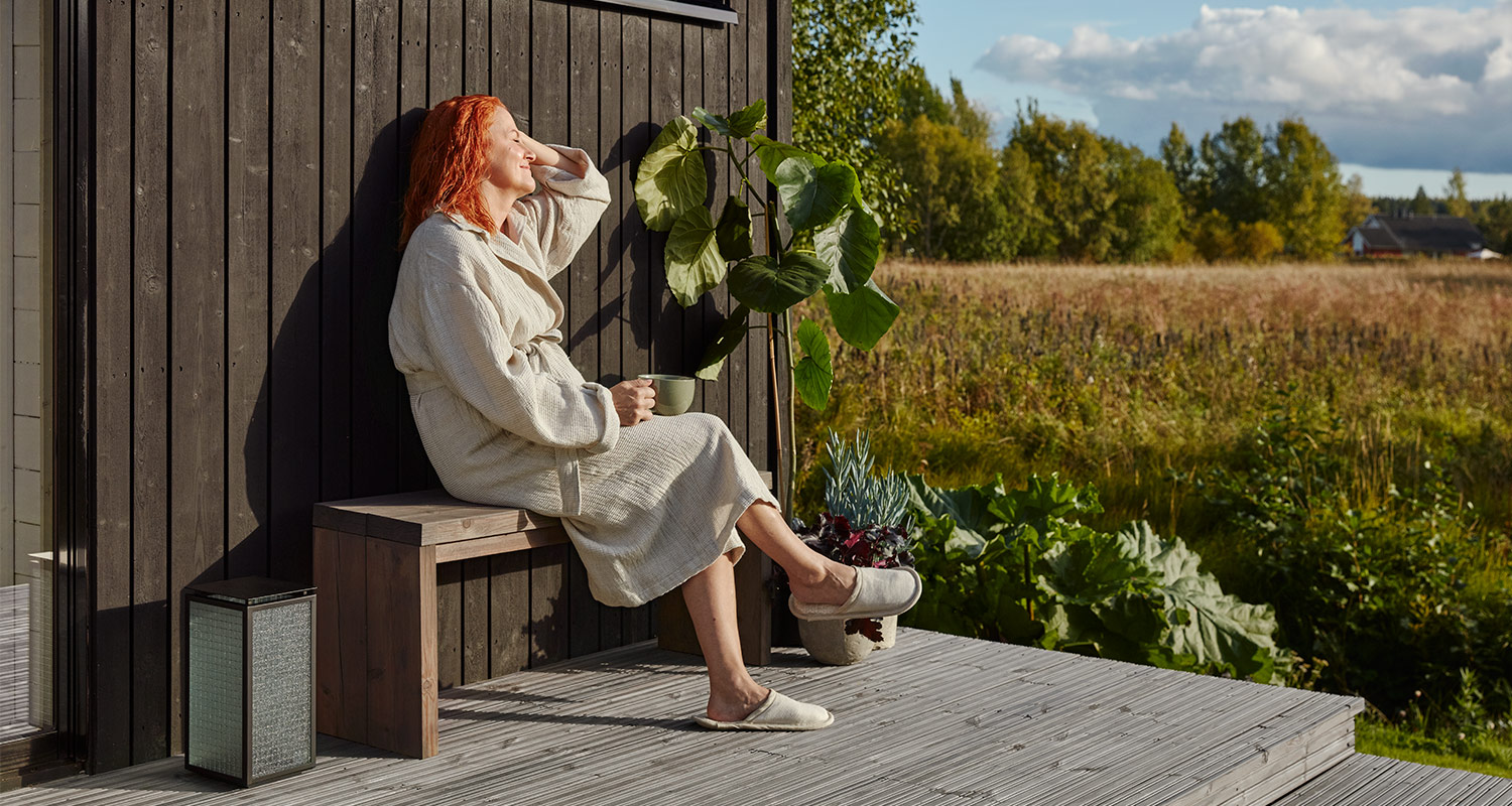 Punatukkainen nainen kylpytakissa pitelee vihreää mukia puupenkillä metsän ja niityn äärellä.