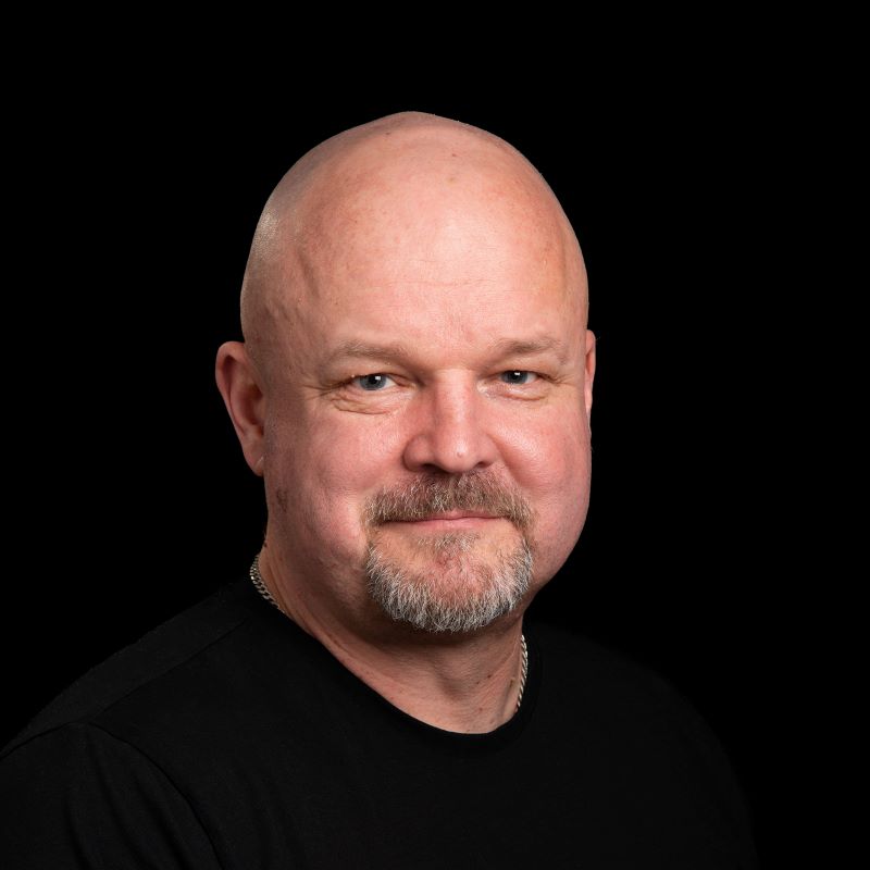 Kalju mies mustassa paidassa ja hopeaketju kaulassa tumman taustan edessä