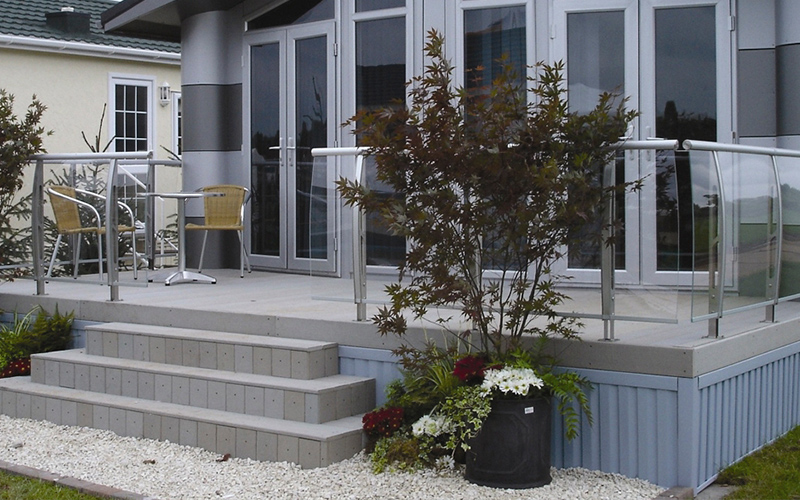 Moderni harmaa terassi lasikaitein, kaksi keltaista tuolia pienellä pöydällä ja kukkia edessä