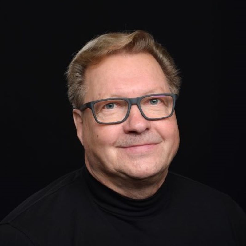 Mies vaaleine hiuksineen mustassa poolopaidassa tummalla studiotaustalla.