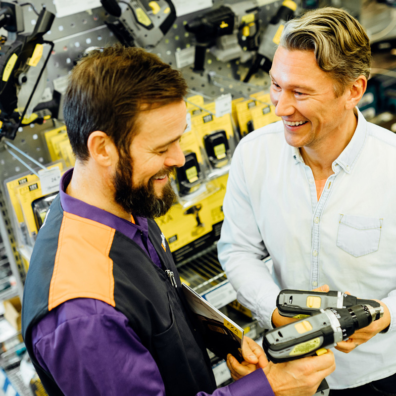 Rautakaupan hyllyjen edessä kaksi miestä vertailee musta-keltaisia porakoneita.