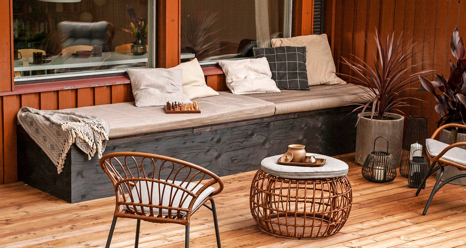 Terassi puulattialla, penkki vaaleine tyynyineen ja shakkilauta, rottinkituolit ja -pöytä