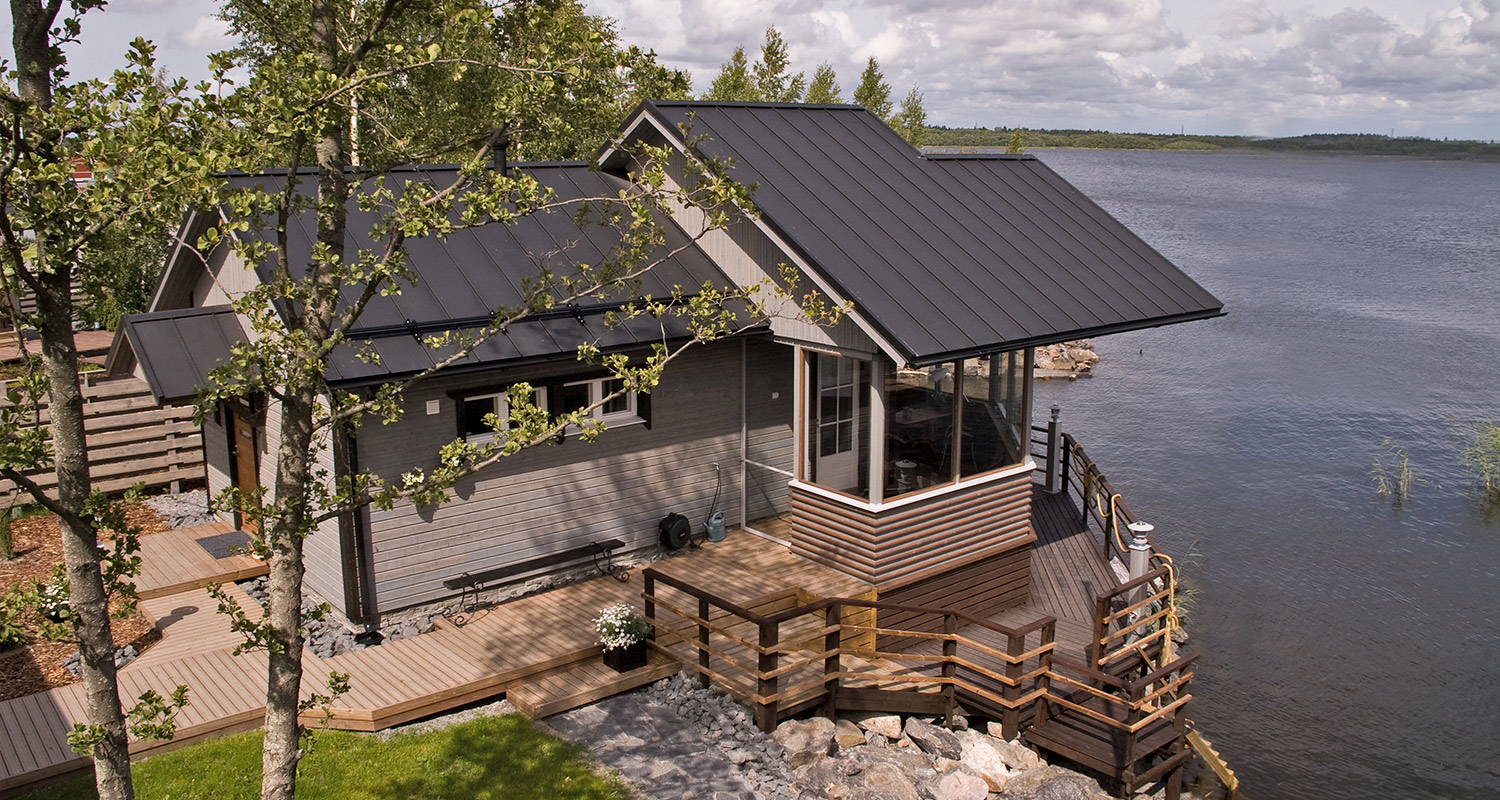 Harmaalankainen mökki mustalla peltikatolla puisella terassilla järven rannalla kesämaisemassa