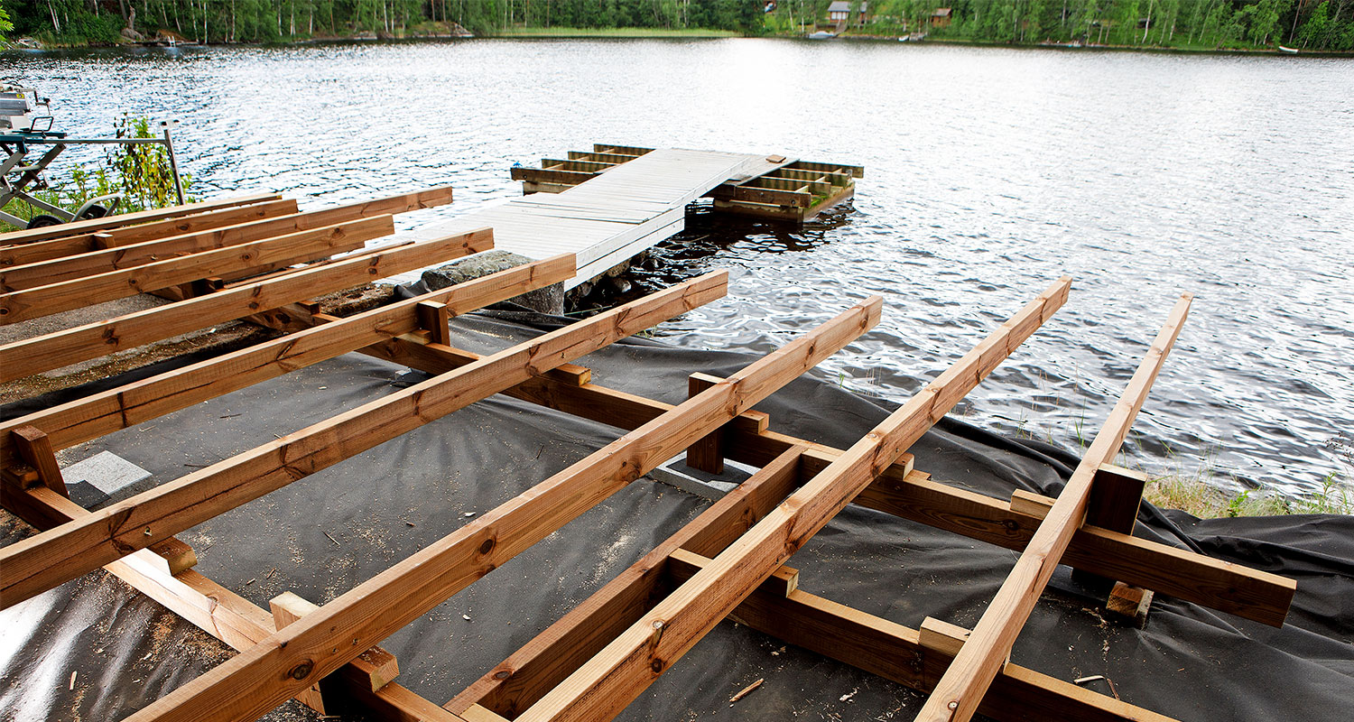 Järvenrannalla työmaa: puurankakehikko laiturin alle, taustalla sininen vesi ja vihreä metsä.