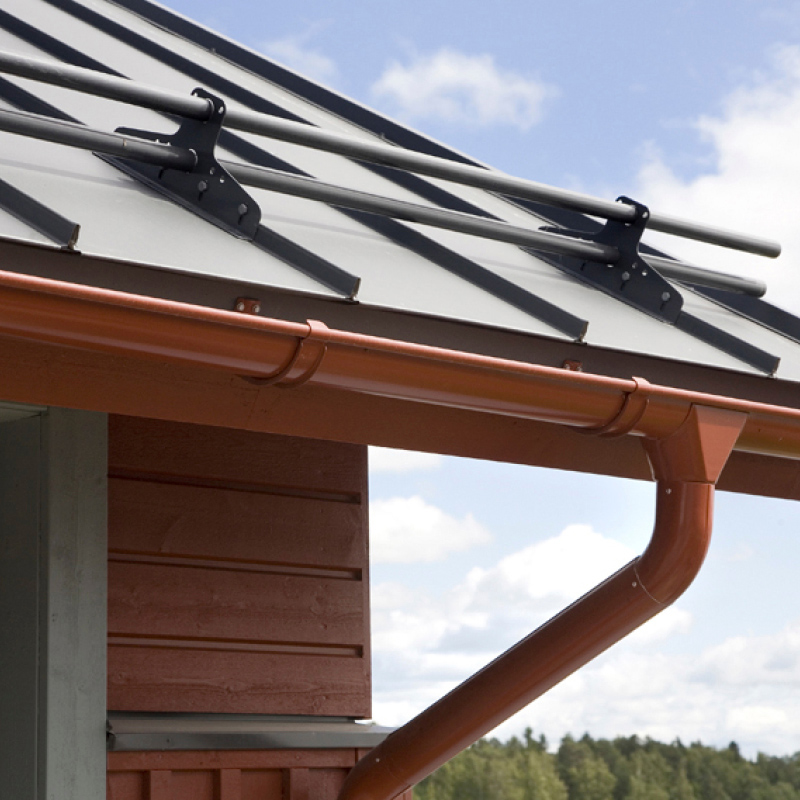 Harmaa metallikatto lumilukoilla ja punainen räystäskouru, taustalla pilvinen taivas ja metsä.