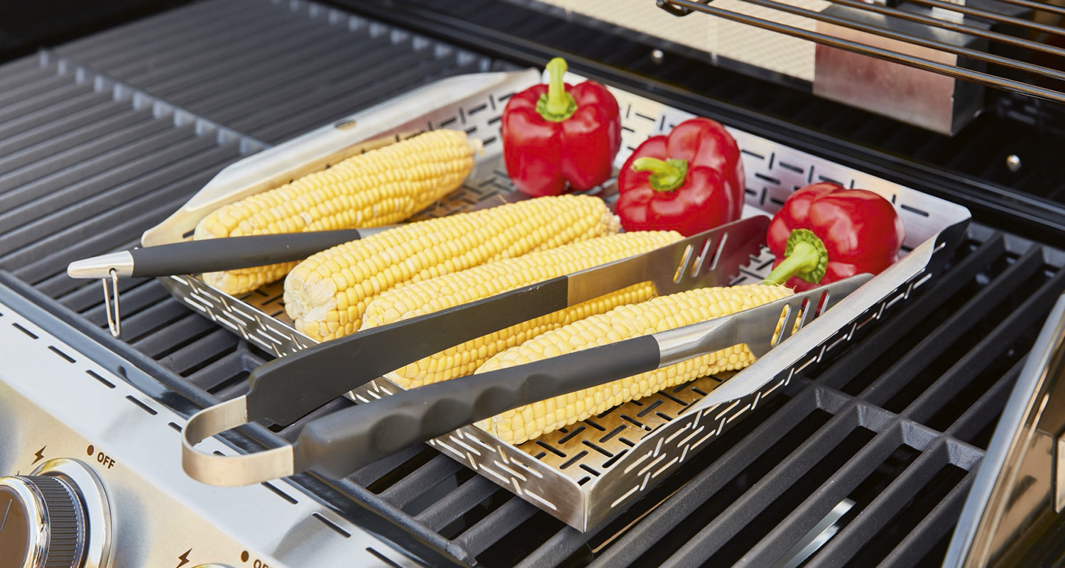 Metallikori grillissä: keltaisia maissintähkiä ja punaisia paprikoita, pihdit päällä.