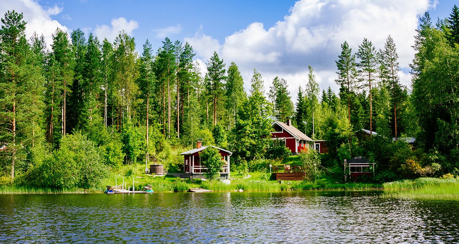 Kaksi punaista mökkiä ja laituri vihreän metsän reunalla kirkkaan sinisen järven äärellä.