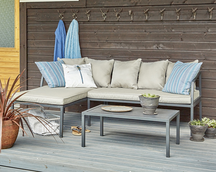 Harmaa L-sohva sinisin ja beigen tyynyin, musta pöytä, ruukkupuut ja seinän peuranpäät.