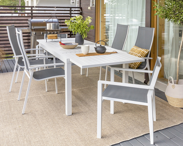 Terassilla valkoinen pöytä harmailla tuoleilla, beige matto, astioita, teekannu ja vihreä kukkaruukku.