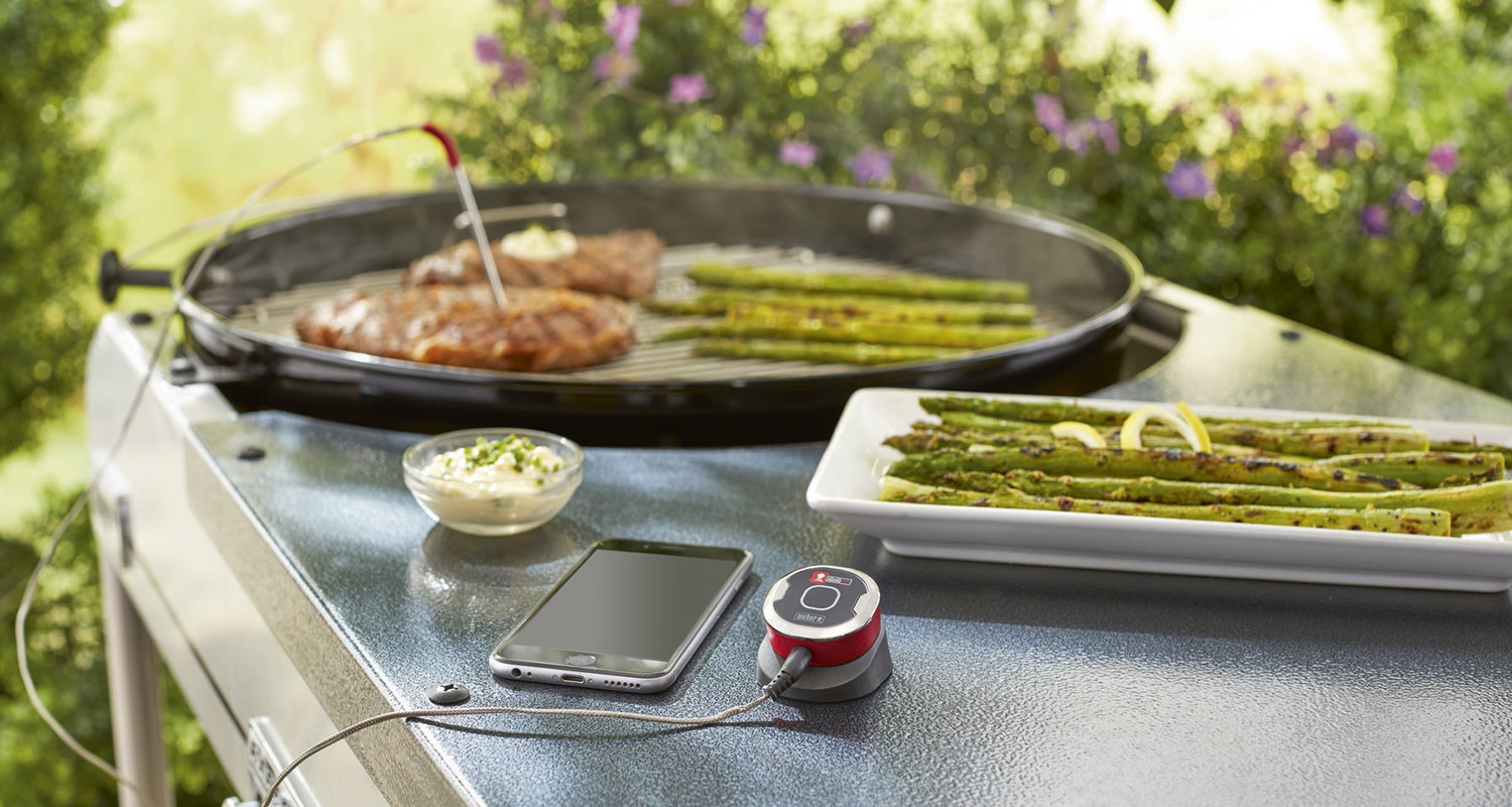 Ulkogrillin sivutasolla älytermostaatti, puhelin ja kastikekulho, grillissä pihvi ja parsaa.