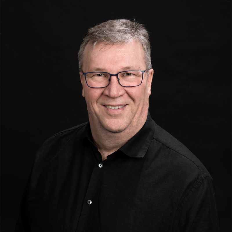 Ylävartalokuva harmaahiuksisesta henkilöstä mustassa kauluspaidassa tummalla taustalla.