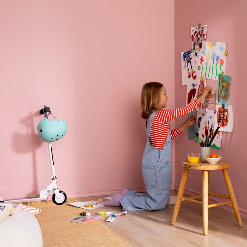 Polvillaan lapsi ripustaa piirustuksia vaaleanpunaiselle seinälle, vieressä potkulauta ja maalit.