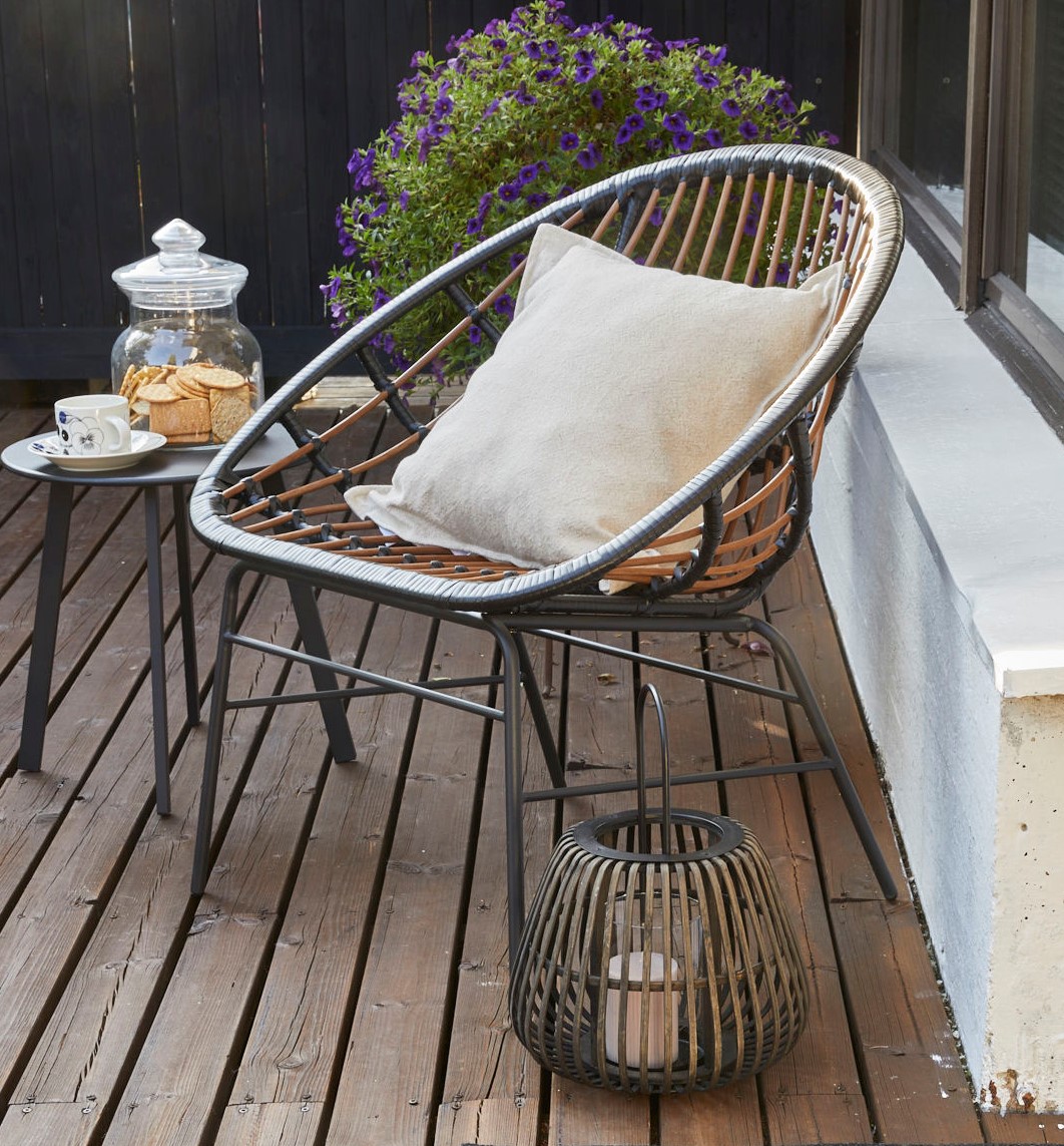 Rottinkituoli vaalealla tyynyllä terassilla, vieressä musta pöytä keksipurkilla ja violetit kukat