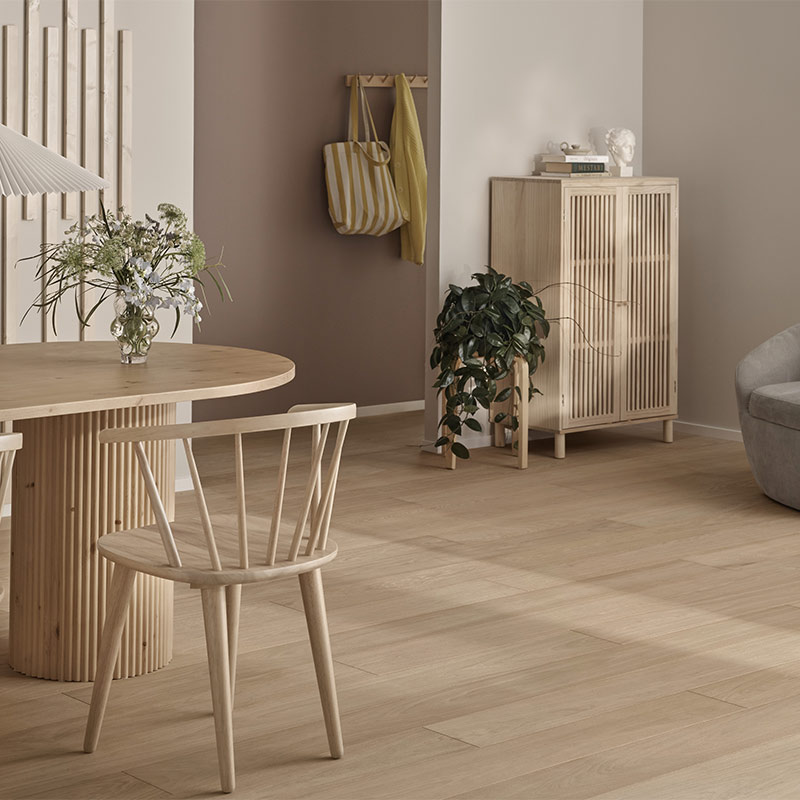 Vaalea sisustus: pyöreä puupöytä kukkavaasilla, puutuoli, lipasto, viherkasvi, keltainen takki