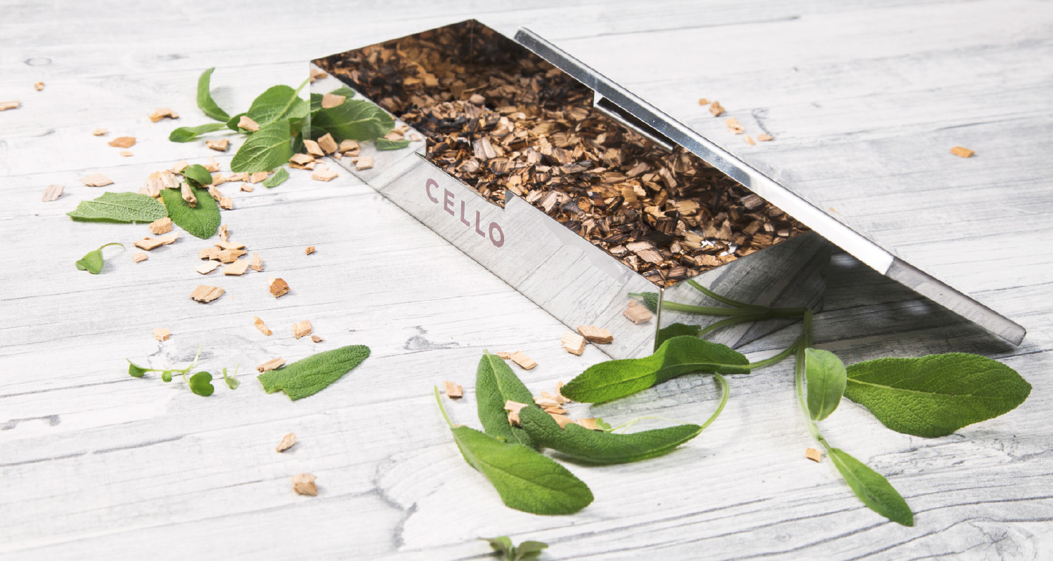 Metallinen Cello-laatikko täynnä puuhaketta, tuoreita vihreitä yrttilehtiä vaalealla puupinnalla