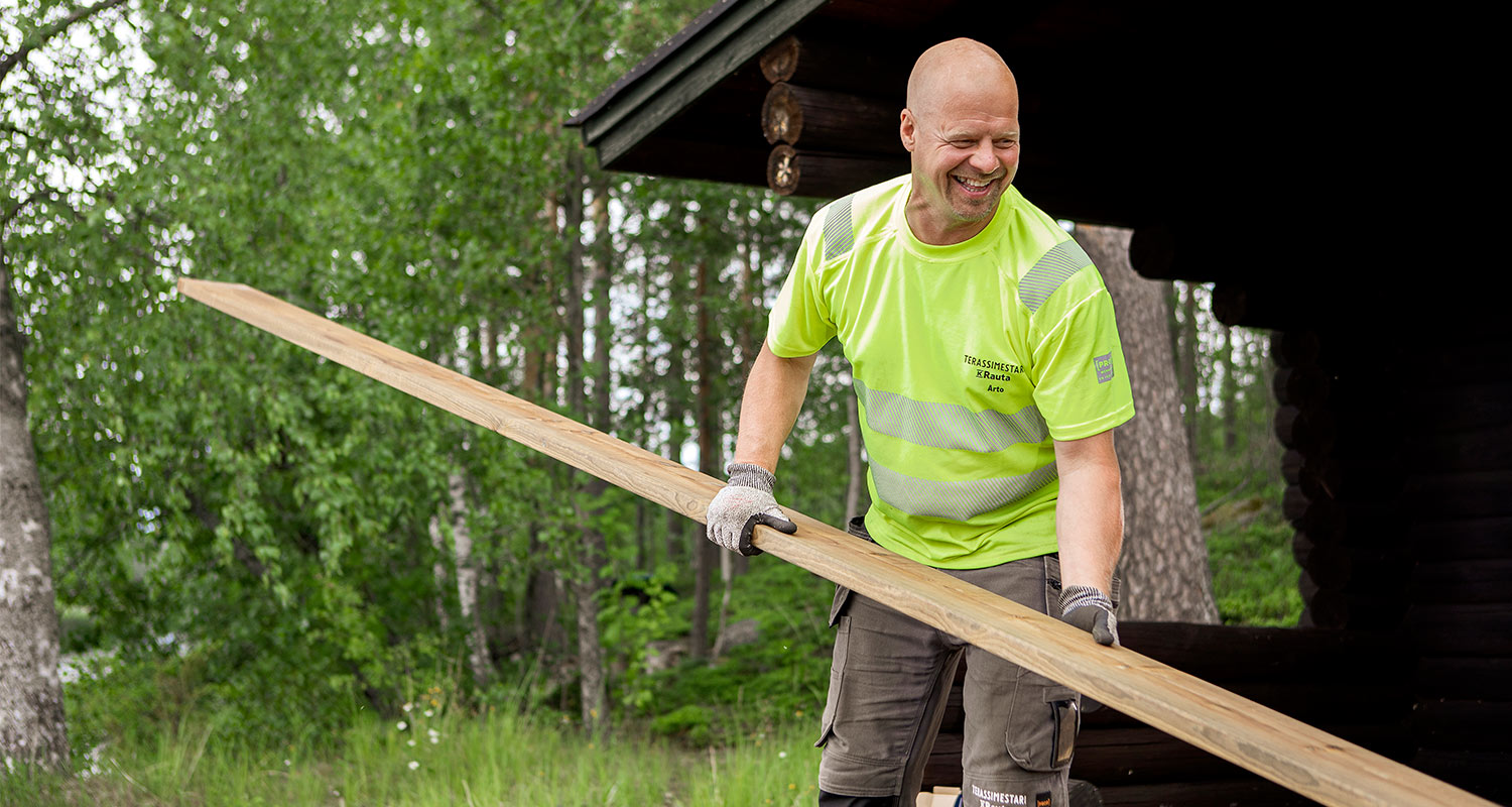 Mies neonvihreässä työpaidassa kantaa pitkää puulistaa metsäisen mökin edustalla.