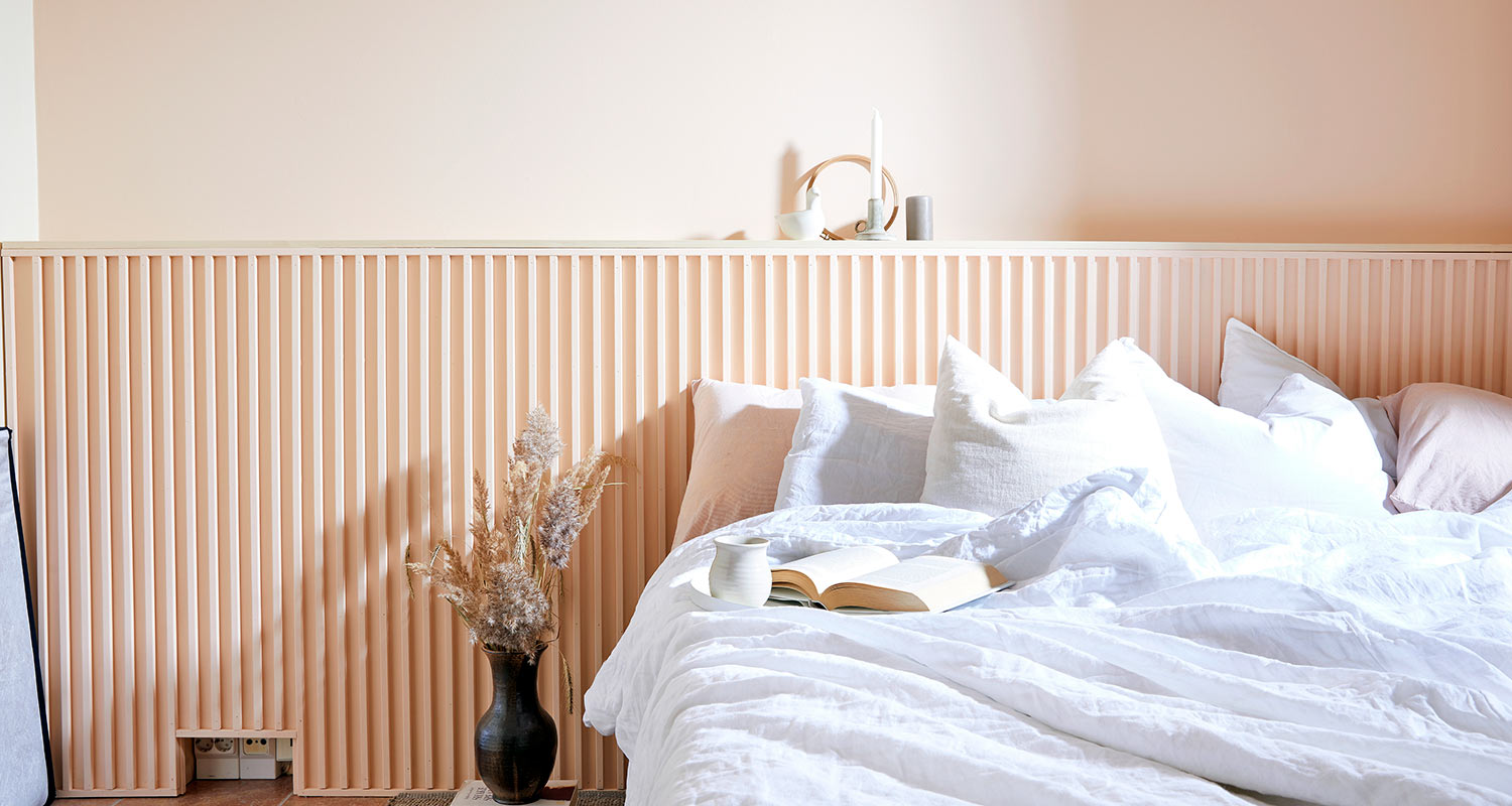 Vaalean persikkapaneeliseinä, valkoinen sänky, avoin kirja ja vaasi kuivakukilla