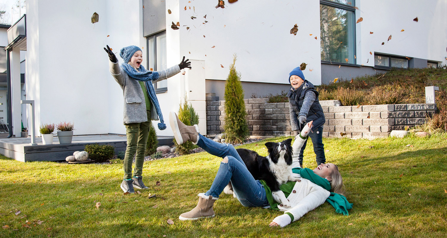 Nainen selällään nurmikolla koira sylissä, lapset heittävät ruskalehtiä valkoisen talon edustalla